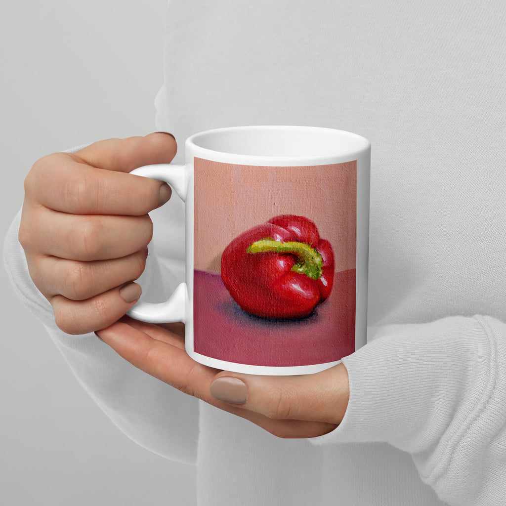 A person in a white long-sleeve top holds a white ceramic mug featuring a printed still life artwork of a red bell pepper on a pink and red background.